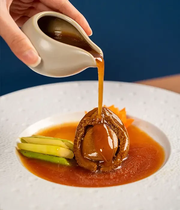 image shows a large abalone sitting in the middle of a large dish with chopped vegetables on the side while a thick brown sauce is being poured on top of it