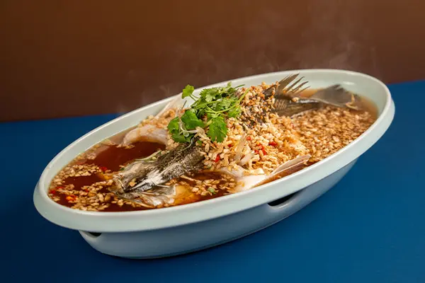 steamed fish in soy sauce with a lot of chopped garlic in a bed of soy sauce topped with cilantro as garnish in an elegant white dish on a blur table top